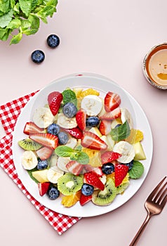 Summer fruit and berry salad with fresh strawberries, blueberries, banana, kiwi, orange and mint, pink background, top view