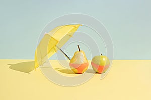 Summer fruit concept. Wet apple and a pear in monokini next to an umbrella isolated on a blue and yellow background.