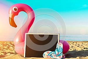 Summer fun beach. Funny pink toy flamingo with blackboard, slippers for text on summer ocean nature beach background in sunny