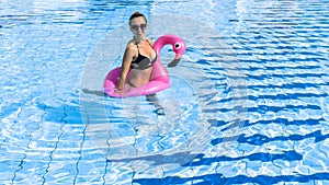 Summer fun beach. Young sexy woman with inflatable pink flamingo in bikini swimsuit, sunglasses float in blue pool water. Spa and