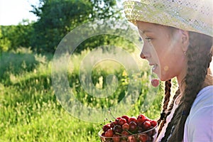 in summer garden young sweet girl in white T-shirt and hat holds ripe cherry berry in her mouth. Delicious and healthy food.