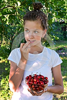 in summer garden young sweet girl in white T-shirt holds a glass vase with ripe cherries and brings one berry to her mouth.