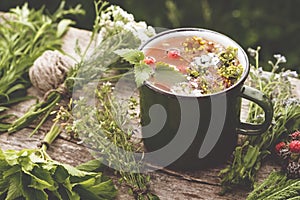 Summer healthy herbal tea in old enameled mug and bunches of healing herbs on wooden board. Herbal medicine.
