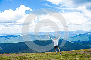 Summer hiking in the mountains. Young tourist man in a cap with hands up on the top of the mountains admires the nature