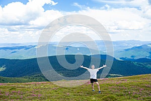 Summer hiking in the mountains. Young tourist man in a cap with hands up on the top of the mountains admires the nature