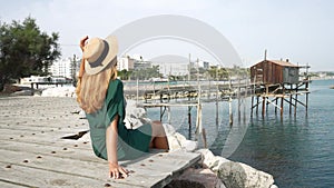 Summer holiday in Italy. Back view of young woman sitting on seafront relaxed with trabucco old fishing machine on Adriatic Sea