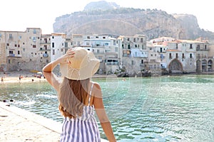 Summer holiday in Italy. Back view of young woman with straw hat and striped dress with Cefalu village on the background, photo
