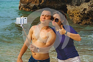 Summer holidays lifestyle portrait of young happy and playful Asian Chinese couple enjoying at the beach taking stick selfie photo