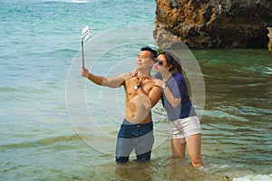 Summer holidays lifestyle portrait of young happy and playful Asian Chinese couple enjoying at the beach taking stick selfie photo