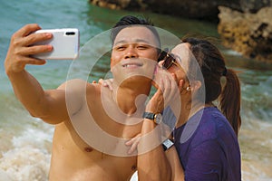 Summer holidays lifestyle portrait of young happy and playful Asian Chinese couple tourist enjoying at the beach taking selfie