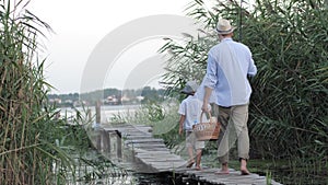 Summer holidays, little boy with fishing rod goes to pier among reeds with father and snack basket to fish on family
