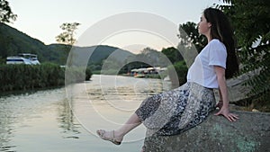 Summer holidays. Young beautiful woman sitting on the pier and swinging her legs, looking at the river. Evening. Side view