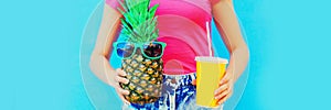 Summer image of close up woman holding cup of juice with pineapple wearing sunglasses on blue background