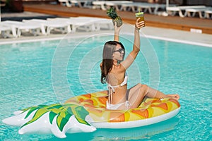 Summer image of stunning suntanned girl swimming on an yellow inflatable pineapple mattress in the pool.