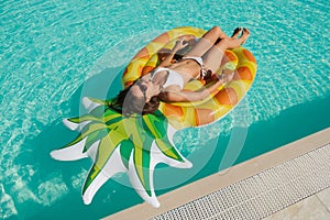 Summer image of stunning suntanned girl swimming on an yellow inflatable pineapple mattress in the pool.