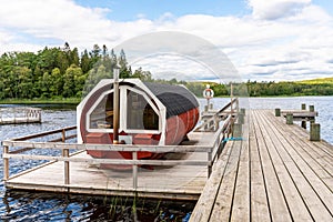 Summer lake nature landscape view of a traditional Scandinavian water floating red wooden sauna spa next to a jetty.