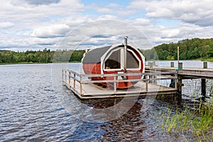 Summer lake nature landscape view of a traditional scandinavian water floating red wooden sauna spa next to a jetty.