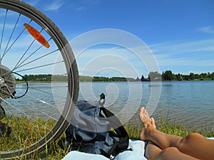 Summer landscape bike whell and barefoot legs girl resting on a lake blue sky background