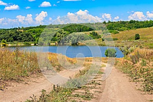 Summer landscape with dirt road to lake and blue sky with white clouds