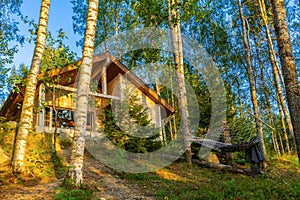 Summer landscape with wooden eco cottage and hammock in forest in evening at sunset. Facade of log cabin among birch trees.