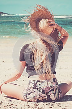 Summer lifestyle blonde long hair young woman sitting at the beach on the sand enjoying blue ocean water in background holding