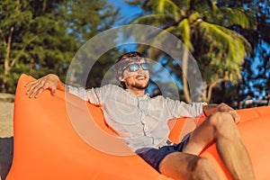 Summer lifestyle portrait of man sitting on the orange inflatable sofa on the beach of tropical island. Relaxing and