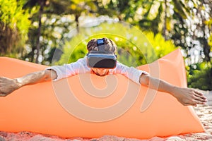 Summer lifestyle portrait of man sitting on the orange inflatable sofa and uses virtual reality headset on the beach of