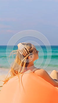 Summer lifestyle portrait of pretty girl sitting on the orange inflatable sofa on the beach of tropical island. Relaxing