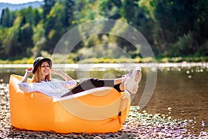 Summer lifestyle portrait of pretty smiling girl lying on yellow inflatable sofa outdoor on river bank. Relaxing
