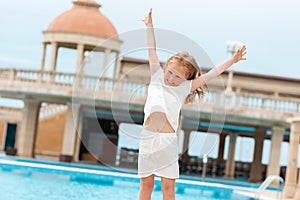Summer. A little Caucasian girl laughing merrily and jumping around the pool. Outdoor. In the background is a complex with a