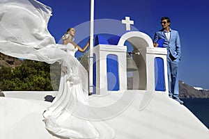 Summer love. Beautiful happy young couple bride and groom in wedding clothes is posing near Church of the Three Bells in Fira on