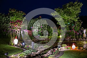 Summer night in garden lightened by illuminated glass objects. Flowering summer garden with pond and artistic glass birds.