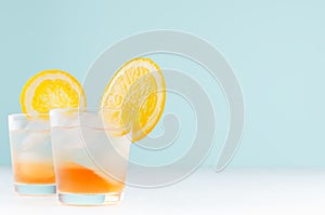 Summer orange lemonade with ice cube, slices oranges in misted glass on elegant pastel green wall, white wood table.