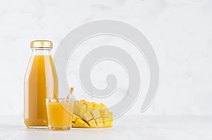 Summer orange pulpy mango in glass bottle mock up with straw, wine glass, fruit pieces on white wood table in light interior.