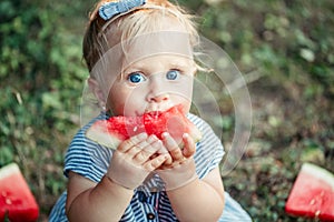 Summer picnic food. Cute Caucasian baby girl eating ripe red watermelon in park. Funny child kid sitting on ground with fresh