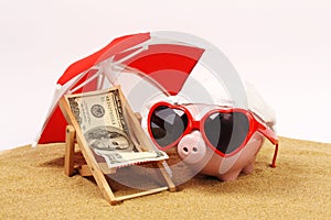 Summer piggy bank with heart sunglasses standing on sand under red and white sunshade next to beach chair with towel from photo