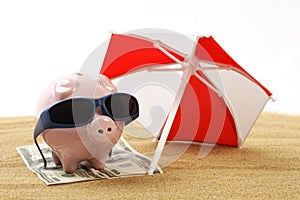 Summer piggy bank standing on towel from greenback hundred dollars with sunglasses on the beach sand under red and white sunshade