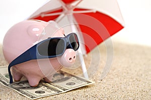 Summer piggy bank standing on towel from greenback hundred dollars with sunglasses on the beach sand unter red and white sunshade photo