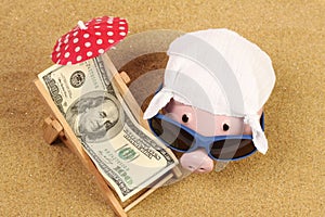 Summer piggy bank with sunglasses and hankie next to beach chair with towel from greenback hundred dollars and red parasol on sand