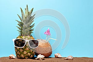 Summer pineapple with sunglasses in sand with coconut drink against a blue background