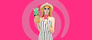 Summer portrait of beautiful woman drinking juice and holding pineapple wearing straw hat, striped jumpsuit, sunglasses on pink