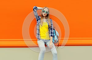 Summer portrait of beautiful young blonde woman posing wearing sunglasses, backpack on orange background