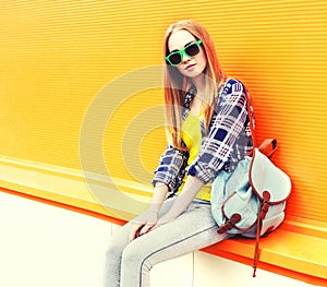 Summer portrait of beautiful young blonde woman wearing sunglasses, backpack on city street on orange background, blank copy space