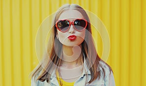 Summer portrait of beautiful young woman blowing lips sending sweet air kiss wearing a red heart shaped sunglasses over yellow