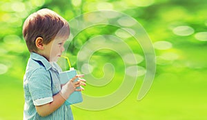 Summer portrait of happy little child drinking fresh juice with straw in the park on a sunny day on blurred green background