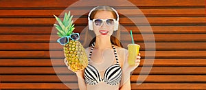 Summer portrait of happy smiling young woman listening to music in headphones with fresh cup of juice, pineapple and sunglasses