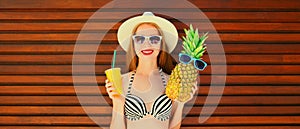 Summer portrait of happy smiling young woman with pineapple and fresh cup of juice wearing summer straw hat on wooden background