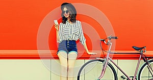 Summer portrait of happy smiling young woman with smartphone and bicycle on city street on red background