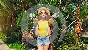 Summer portrait of happy young woman with fresh pineapple wearing straw hat, sunglasses on the beach with palm tree background