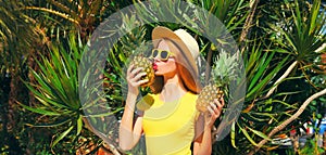 Summer portrait of happy young woman kissing fresh pineapple wearing straw hat, sunglasses on the beach with palm tree background